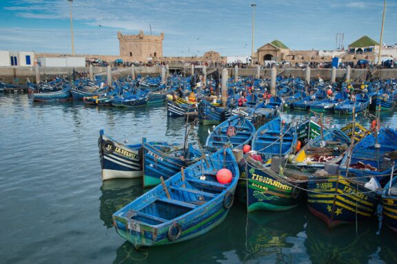 A bustling harbor scene in Essaouira, Morocco with numerous blue fishing boats moored closely together. A stone fortress overlooks the scene in the background.