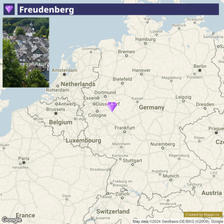 A map highlighting Freudenberg, Germany with a location marker, and an inset photo in the upper left corner showing a close-up view of traditional half-timbered houses in Freudenberg. Major nearby cities such as Cologne and Dortmund are visible on the map, as well as the borders of neighboring countries: Netherlands, Belgium, Luxembourg, France, and Switzerland.