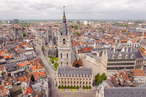 Aerial view of the historic Ghent city center in Belgium with dense medieval buildings, featuring a prominent Gothic cathedral with a tall belfry adorned with a golden statue, surrounded by a large square and various architectural styles.