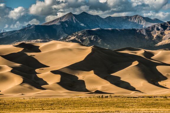 Great Sand Dunes National Park Colorado featuring large sand dunes with dramatic shadows in the foreground, backed by a towering mountain range under a cloudy sky.