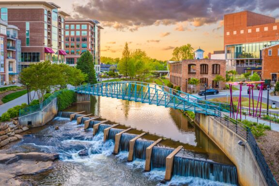 A picturesque urban sunset scene featuring a pedestrian bridge over a river with cascading waterfalls, surrounded by mixed-use buildings, lush greenery, and a clear sky with clouds reflecting the warm hues of the setting sun.