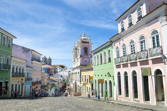 A colorful historic district with colonial architecture, cobblestone streets, and people walking under a clear blue sky. A prominent baroque church with twin towers stands in the background.