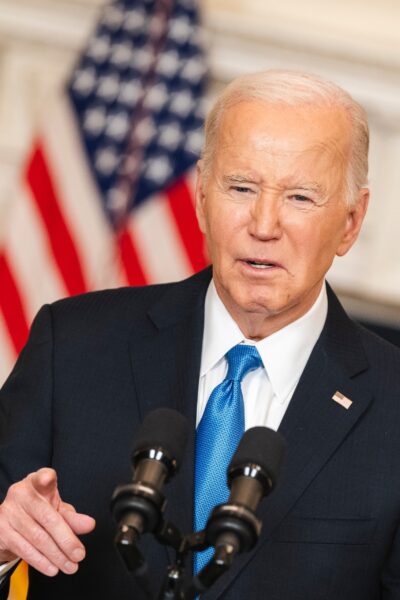 Joe Biden in a dark suit and blue tie speaking into microphones with the American flag in the background.