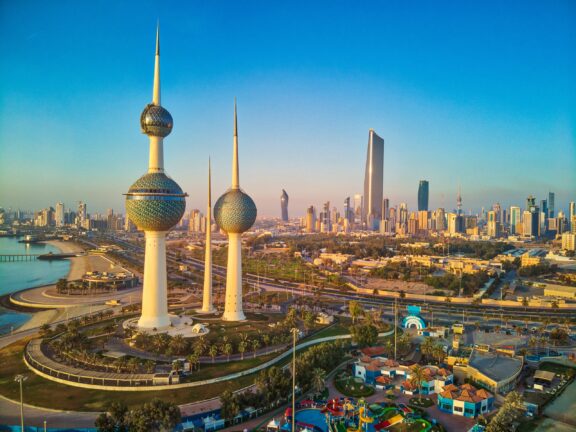 Aerial view of Kuwait City skyline at sunset with the iconic Kuwait Towers in the foreground and modern skyscrapers in the background.