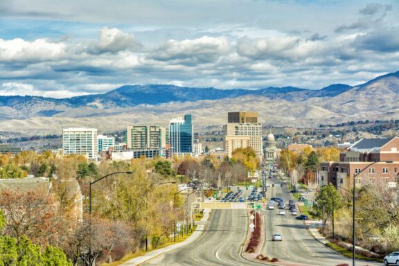 A view of a cityscape with modern buildings, a tree-lined street leading into the downtown area, with a backdrop of rolling hills and a cloudy sky.