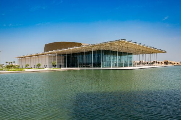 Modern waterfront building with a large glass facade and a unique, angled roof structure against a clear blue sky.