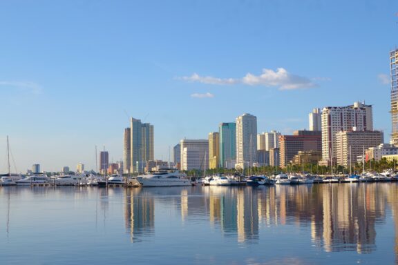 A scenic view of a city skyline with various high-rise buildings reflecting on the calm water of a harbor filled with moored boats and yachts under a clear blue sky.