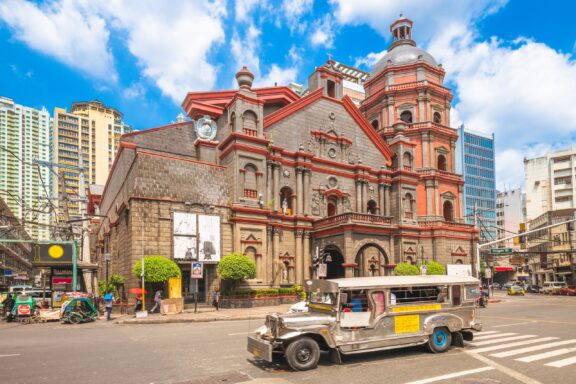 A historic stone church with a red dome and accents in Manila, flanked by modern skyscrapers, with a jeepney in the foreground on a sunny day with blue skies.