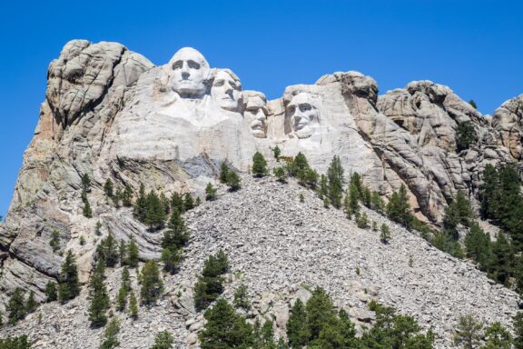 Mount Rushmore National Memorial depicting the colossal carved faces of four former U.S. presidents surrounded by pine trees and a blue sky.