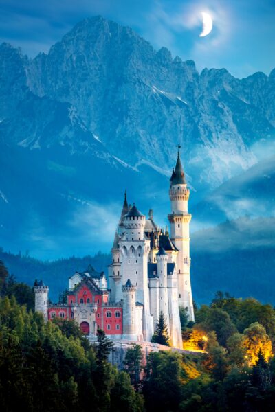 A fairytale-like castle with multiple spires illuminated by a warm light, set against a backdrop of craggy mountains and a twilight sky with a crescent moon.