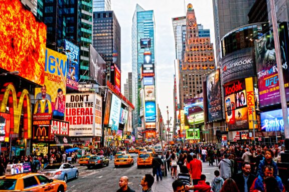 A bustling Times Square with illuminated billboards and advertisements, heavy traffic with yellow taxis, and crowded sidewalks with pedestrians.