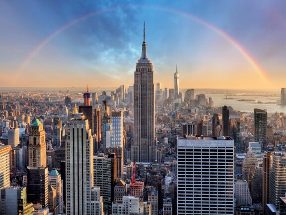 A panoramic view of New York City skyline at sunset with the Empire State Building in the foreground and a double rainbow in the background.