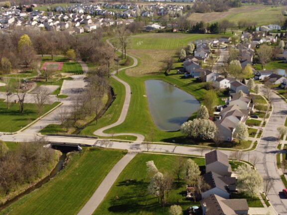 Aerial view of a suburban neighborhood with houses, a small pond, and a park with a tennis court on a sunny day.