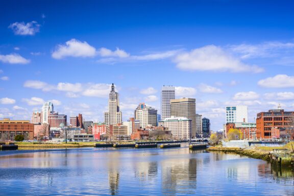 Downtown skyline of a city with various buildings reflected in the calm water of a river on a sunny day with scattered clouds.