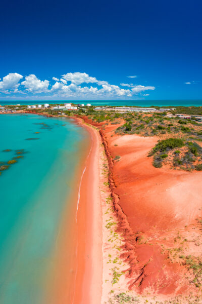 Aerial view of a turquoise shoreline with a distinct red sandy beach, bordered by green vegetation and buildings against a backdrop of fluffy clouds in a blue sky.