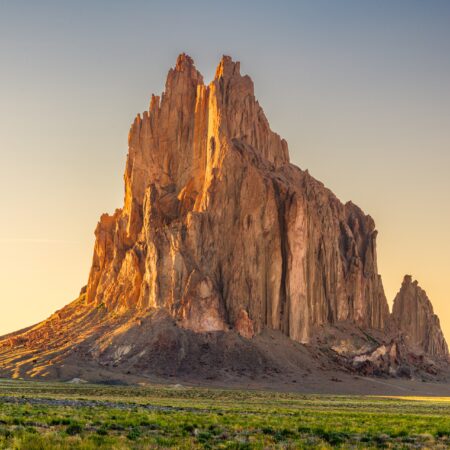 An imposing rock formation bathed in warm sunlight during golden hour, with sharp peaks rising against a clear sky and a foreground of green vegetation.