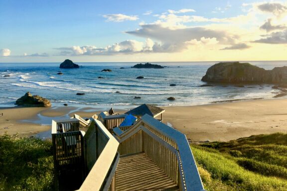 A wooden staircase leads down to a serene beach with scattered rocks and islets near the shore, under a partly cloudy sky at sunset in Coquille Point, Bandon, Oregon.