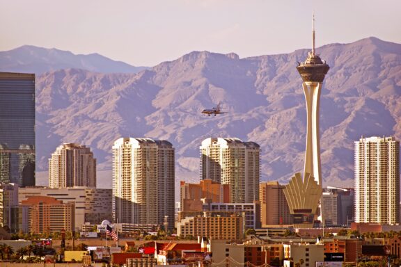 A view of Las Vegas skyline with the Stratosphere Tower prominent against the backdrop of mountain ranges and a clear sky, and an airplane flying in the distance.