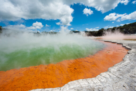 A vibrant geothermal hot spring with orange microbial mats, steam rising above the turquoise water, under a blue sky with puffy white clouds.