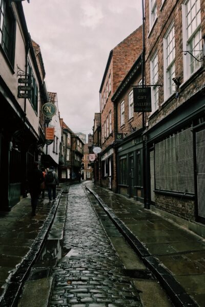 Narrow cobblestone street with pedestrians and historical buildings showing shop signs, wet from rain, in an old town setting.