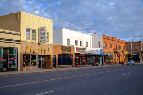 A row of small commercial buildings including a florist under a blue sky with scattered clouds. A street lies in the foreground with a lone car parked on the side.