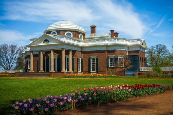 A neoclassical red brick mansion with white columns and trim, dome, and balconies, surrounded by lush gardens with blooming tulips under a clear blue sky.