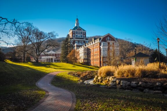 A winding brick pathway leading up to a large, historic multi-story building with a clock tower, set against a backdrop of hills and a clear blue sky.