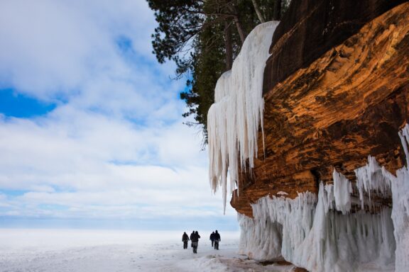 A group of people walking on a snow-covered ground near a large cliff with striking icicles hanging down. A clear blue sky with a few clouds is visible above.