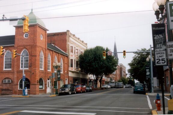 A view of a small town street with historic brick buildings, parked cars, traffic signals, and a church spire in the distance under an overcast sky. Signs indicating a speed limit of 25 and welcome banners are visible on the right.