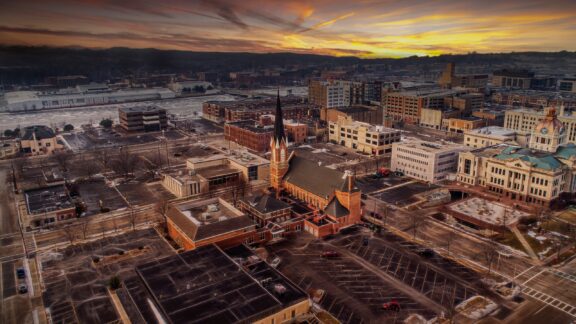 Aerial view of a cityscape at sunset with buildings, streets, parking lots, and a prominent church with a tall spire, against a backdrop of mountains and a vibrant sky.