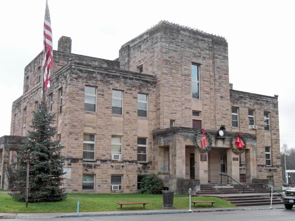 A stone building with a central staircase leading up to a double-door entrance flanked by windows with Christmas wreaths, a Christmas tree to the left, and an American flag flying above.