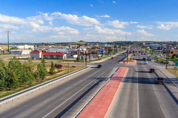 A wide boulevard with multiple lanes of traffic and a red bus lane, leading through a commercial area with various shops and signs under a clear blue sky.