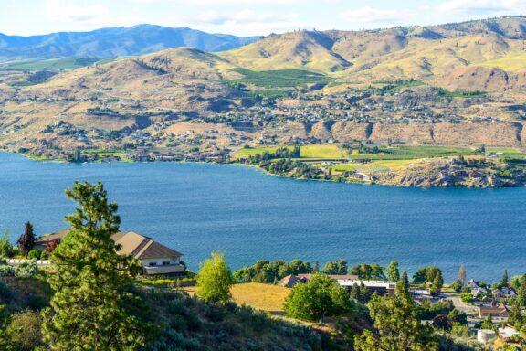 A scenic view of a lakefront community nestled among rolling hills with houses scattered along the shore and greenery in the foreground.