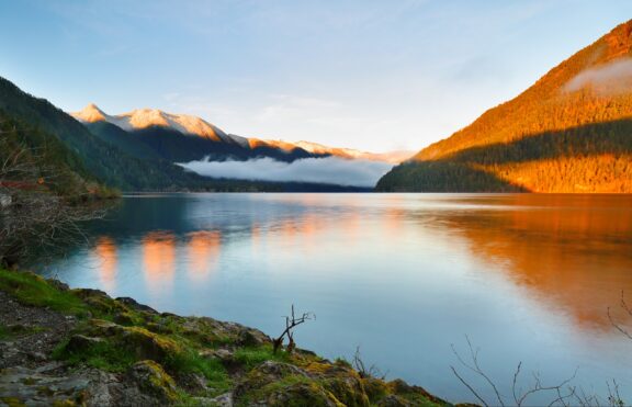 A serene lake at sunset with surrounding mountains partially lit by the golden sun, reflected in the water, and low-hanging clouds nestled in the valleys.