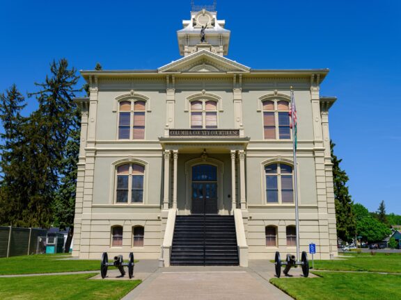 A front view of the historic Columbia County Courthouse with a clear blue sky, featuring a clock tower, American flag, and cannons on the lawn.