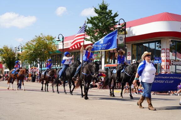 A group of equestrians in blue and white outfits carrying American flags and a "Morgan Horse Club" banner while participating in a parade on a sunny day with spectators watching from the sidewalks.