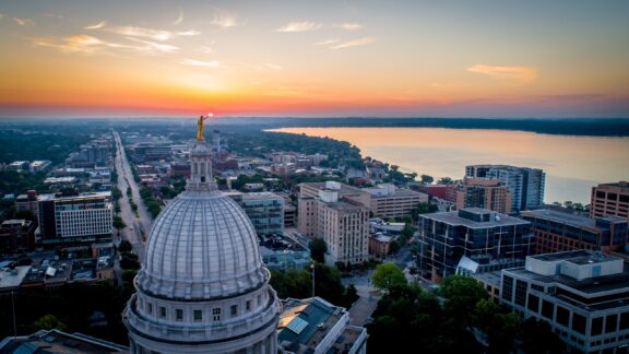 Aerial view of a city at sunset with a domed capitol building in the foreground and a vast river in the background under a sky with scattered clouds.