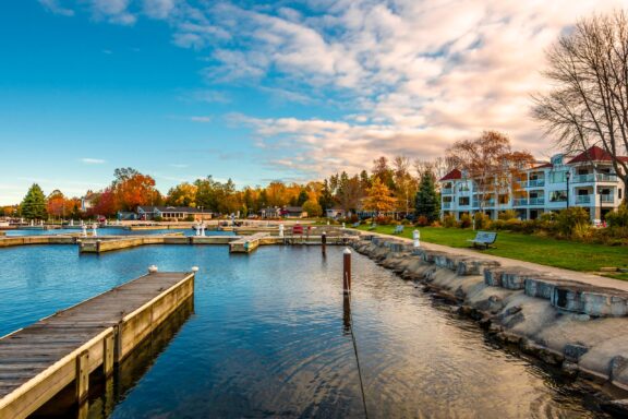 A picturesque marina with empty wooden docks, calm blue water, surrounded by colorful autumn trees and modern buildings under a cloudy sky.