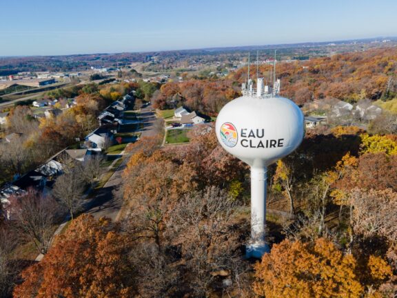 Aerial view of a water tower with "EAU CLAIRE" painted on it, surrounded by autumn-colored trees with a residential area in the background.