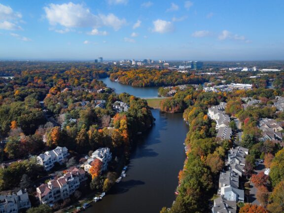 Aerial view of a suburban area with colorful autumn foliage, a river winding through the landscape, residential houses along its banks, and a city skyline in the distance against a partly cloudy blue sky.