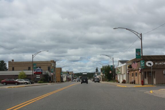 A view of a small town street with vehicles, lined by buildings with American flags, under an overcast sky.