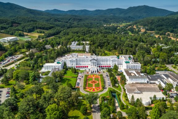 Aerial view of a large, white multi-story resort hotel with red-roofed entrances and a finely manicured garden in the foreground, surrounded by lush trees with rolling hills in the background under a clear sky.