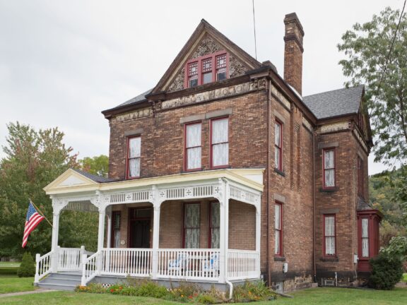 A two-story brick Victorian house with decorative trim, a covered front porch with white railings, and an American flag displayed to the left.