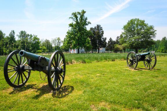 Two antique cannons on display in a green field with trees and white buildings in the background under a clear blue sky.