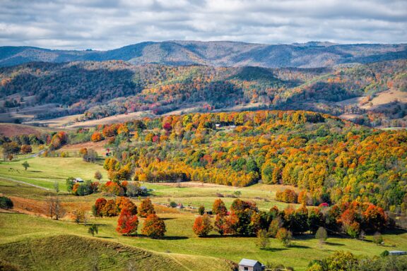 A vibrant autumn landscape featuring rolling hills with colorful foliage, farmhouses, and a shadow-casting cloud pattern overhead.