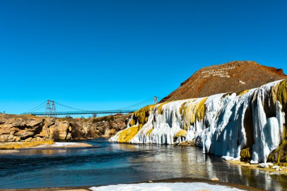 Alt text: A vibrant landscape featuring a partially frozen waterfall with icicles, a flowing river, a suspension bridge in the distance, and a clear blue sky, with a hill prominently displaying white lettering on its side.