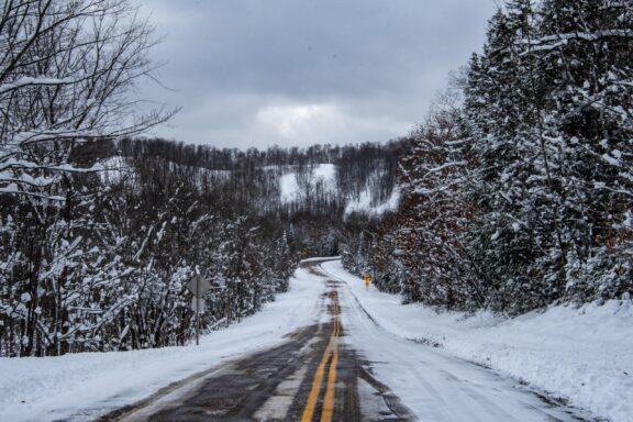 A snowy road meandering through a dense forest with snow-covered trees on an overcast day.