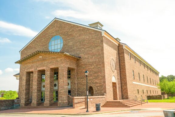 A large brick building with a semi-circular window at the peak, multiple arched doorways, and a series of steps leading to the entrance, set against a clear sky with some greenery on the sides.