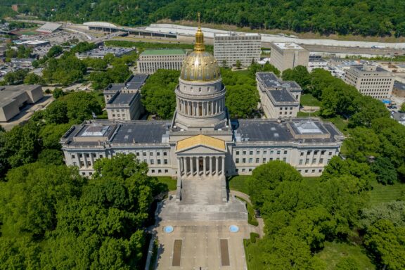 Aerial view of a large neoclassical state capitol building with a golden dome, surrounded by trees and set against an urban backdrop.