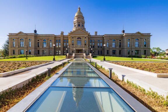 A majestic historical building with a central dome and clock, flanked by two symmetrical wings with a clear blue sky in the background. In the foreground, there is a reflecting pool leading up to the building's entrance with landscaped garden beds on either side.